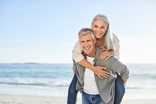 Portrait of smiling man piggybacking woman. Mature couple is enjoying at beach on sunny day. They are on vacation.
