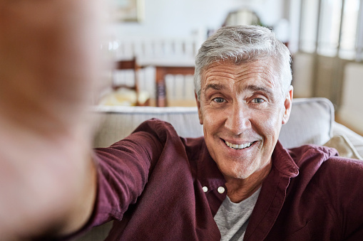 Close-up of smiling mature man taking selfie. Male is spending leisure time at home during coronavirus lockdown. He is in living room.