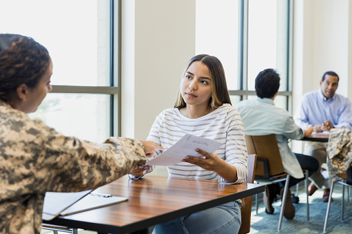 A mid adult female recruiter points to the paperwork the young adult woman is holding. As she explains the application process, the young woman listens attentively.