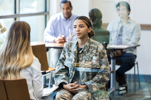 The young adult female soldier has an angry look on her face as she sits with her hands clasped in her lap.  She is listening to the unrecognizable female counselor.