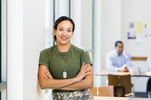 A mid adult female soldier leans against the wall with her arms crossed.  She is taking a break from work to smile for the camera.