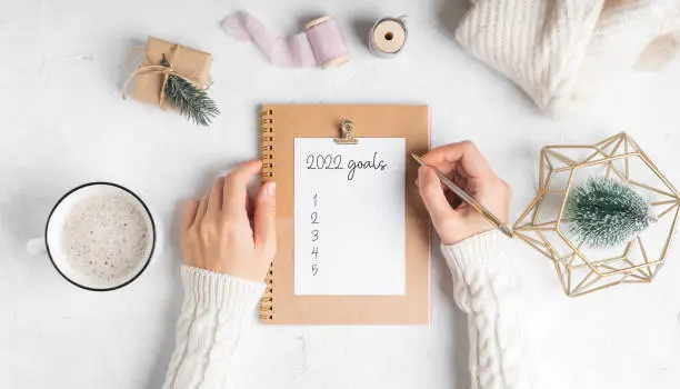 Desktop of woman hands with blank notepad writing 2022 goals text . Flat lay of white working table background with cup of coffee and Christmas decoration. Top view mock up