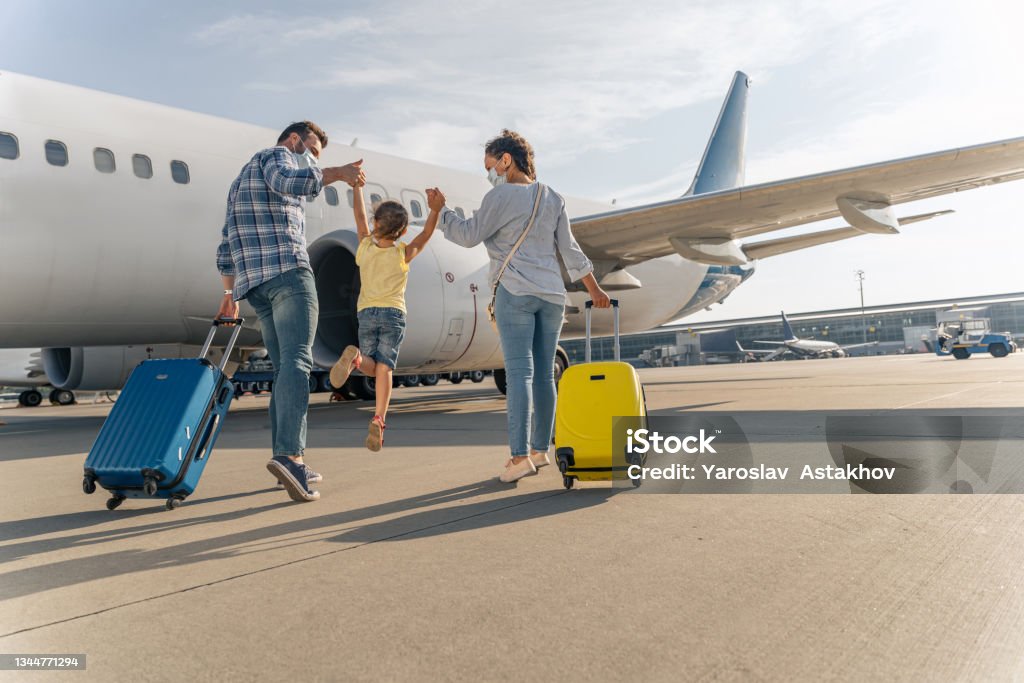 Happy family in masks enjoying travel together Back view of parents holding the hands of the child and going with suitcases to board the plane Travel Stock Photo