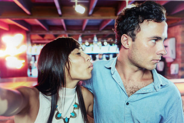 Girlfriend puckers up as she takes a selfie, but reluctant boyfriend turns away Young woman extends her arms as she takes a selfie, but boyfriend doesn't want to take part. vex nightclub stock pictures, royalty-free photos & images