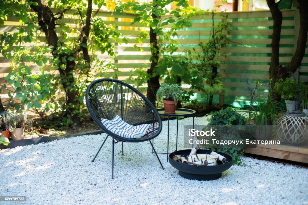 Front veranda of house with black Acapulco armchairs, coffee table and plants pots. Metal black fireplace bowl in garden on back yard patio. Outdoor patio furniture on pebbles in a front garden. Back Yard Stock Photo