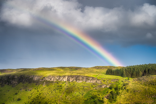 Closeup on vivid rainbow arching over hills and forests covered in patches of sunlight, Glenariff Forest Park, County Antrim, Northern Ireland
