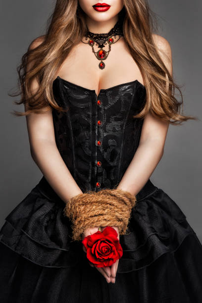 sexy junge frau in schwarzem korsettkleid mit seil gebunden hände, die rote rosenblume halten. art fantasy hexe gothic mädchenporträt mit roten lippen im historischen kleid über dunkelgrauem hintergrund - korsett stock-fotos und bilder