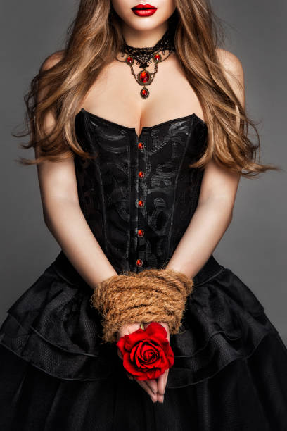 sexy young woman in black corset dress with rope tied hands holding red rose flower. art fantasy witch gothic girl portrait with red lips in historical gown over dark gray background - bustiers imagens e fotografias de stock