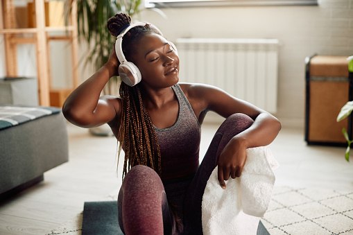 Young African American athletic woman enjoying in music over headphones while taking a break during home workout.