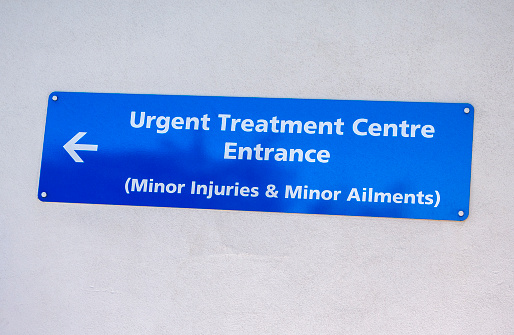 A blue sign directing patients to the Urgent Treatment Centre Entrance for Minor Injuries & Minor Ailments on a wall outside a hospital building.