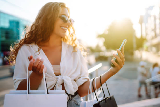 istock Young woman using phone, holding shopper bags, standing outdoors, choosing and ordering clothes and presents. ​ 1344805410