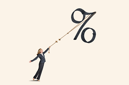 A woman attempts to rein in a large interest rate symbol with a rope that is frayed and about to break.