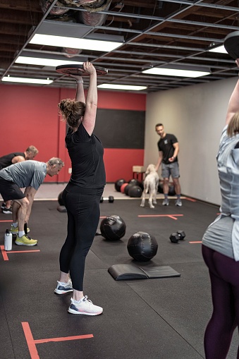 A group of men and woman lift barbell plate above their head during a cross training workout. They are attending a group fitness class focused on strength and functional fitness. The gym owner is coaching the class and is visible in the background petting his obedient dog. Selective focus on a woman near the center of the frame.