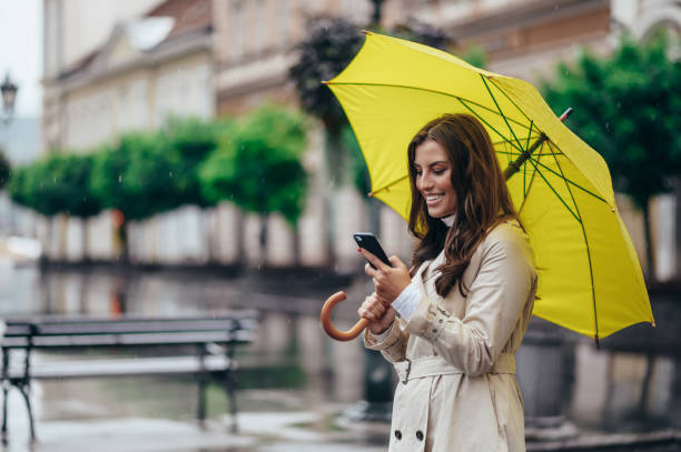 kobieta korzystająca ze smartfona i trzymająca żółty parasol podczas pobytu w mieście - umbrella women storm yellow zdjęcia i obrazy z banku zdjęć