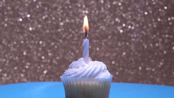 istock Cupcake with single birthday candle burning and sugar sprinkles icing on gold background. Delicious muffin decoration. Homemade vanilla cup cake with buttercream frosting. Shallow depth of field. 1344817445