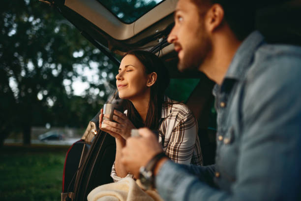 Young couple in love sitting in car drinking coffee during sunset Young couple in love sitting in car open trunk drinking coffee couple driving at night stock pictures, royalty-free photos & images