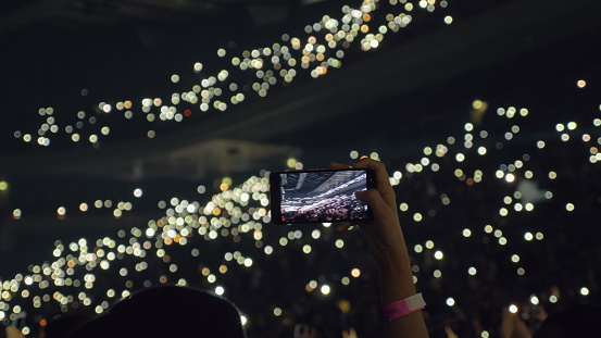 Woman taking mobile video of crowded concert hall. People with lights in the darkness