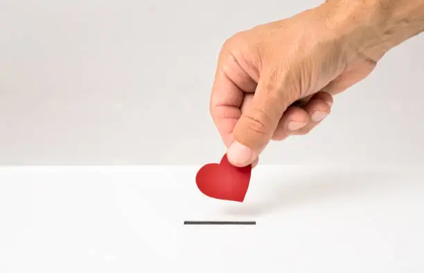 Photo of Red heart symbol is put by person's hand into slot of white donation box