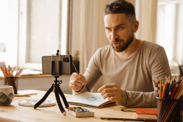 Portrait of Mid-adult artist male. Dark haired man smiling happy making online art class for students at studio. Holding watercolor drawing in his hand. Concept zoom Remote learning. Real emotions. retrato del artista de mediana edad masculino. hombre de cabello oscuro sonriendo feliz haciendo clase de arte en línea para estudiantes en el estudio. sosteniendo un dibujo de acuarela en la mano. concepto zoom aprendizaje remoto. emociones reales
