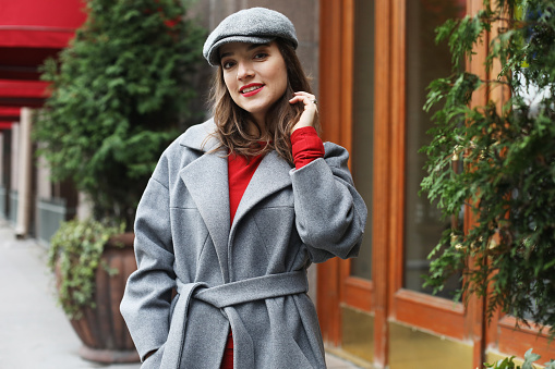 Lifestyle and people concept: young stylish pretty woman wearing red dress, grey coat and hat posing in the city streets.