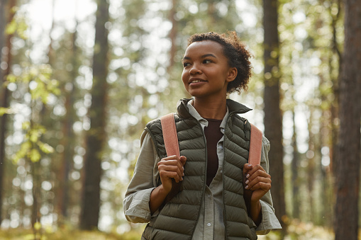 Waist up portrait of young African-American woman with backpack enjoying hiking in forest lit by sunlight, copy space