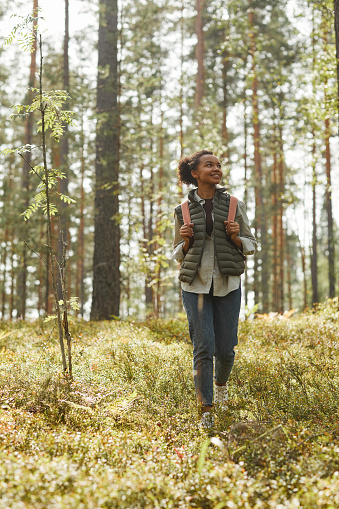 Vertical full length portrait of young African-American woman with backpack enjoying hiking in forest lit by sunlight