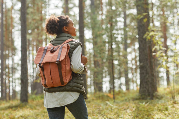 Young Woman with Backpack Outdoors Back view portrait of young African-American woman with backpack enjoying hiking in forest lit by sunlight, copy space exploration stock pictures, royalty-free photos & images