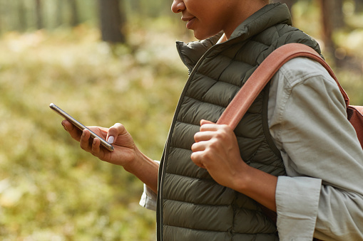 Cropped side view shot of young African-American woman using smartphone while enjoying hiking in forest, copy space