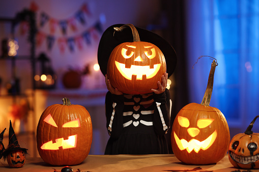 All hallows eve. Little girl child in witch hat and skeleton costume holding glowing jack o lantern carved pumpkin in front of her face, standing behind table with Halloween decorations in dark room