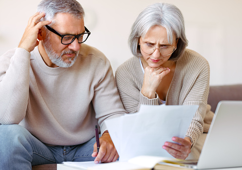 Worried senior family couple reading financial documents or notification letter with bad news from bank while paying bills online on laptop, retired husband and wife having money problems