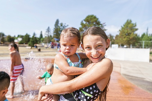Cute elementary age girl holds her baby sister and smiles directly at the camera while playing at a splash pad water park on a hot and sunny summer day.