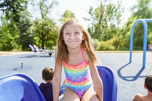 A cute blonde girl wearing a rainbow swimsuit makes a silly happy face while playing on the playground on a hot, summer day.