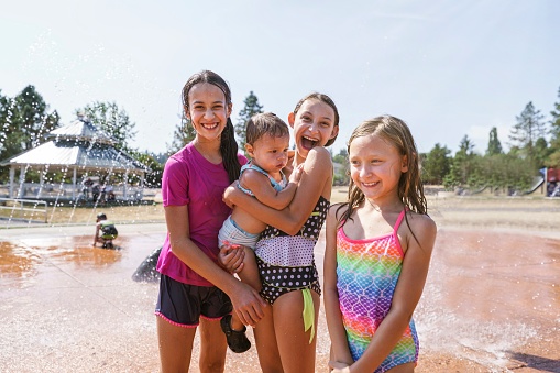 Mixed race group of girls smile and pose for the camera while playing together in the fountain at a splash pad water park on a sunny and hot summer day. The kids vary in age and the girl in the center of the frame is holding an 18 month old toddler. Summer, happiness, cousins, and friendship concepts