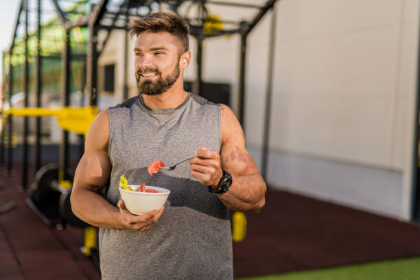 Happy young bodybuilder eating a vegetable salad. Smiling young male athlete eating a vegetable salad at the gym. eating body building muscular build vegetable stock pictures, royalty-free photos & images