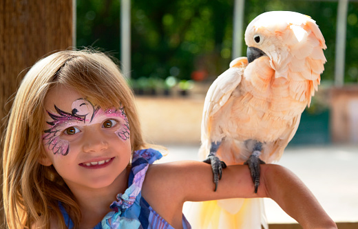 Young girl with face painted in bird style, holds cockatoo on arm