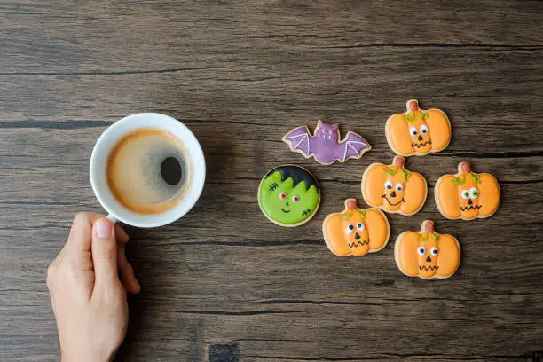 Photo of hand holding coffee cup during eating funny Halloween Cookies. Happy Halloween day, Trick or Threat, Hello October, fall autumn, Traditional, party and holiday concept