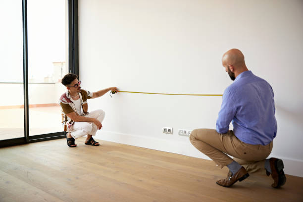 Mid Adult Couple Measuring Wall During Real Estate Showing Full length view of men crouching and holding tape measure to wall of unfurnished living room as they gather property details. measuring a room stock pictures, royalty-free photos & images