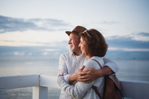 Portrait of a happy senior couple in love hugging outdoors on pier by sea, looking at view, summer holiday.
