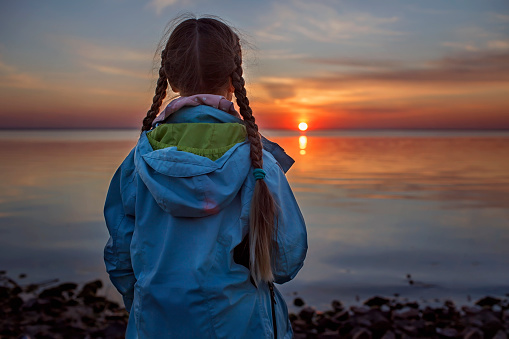 A cute girl with two pigtails in a coat stands on the seashore and enjoys the sunset, escape to nature, digital detox, time to be alone. Autumn outside lifestyle