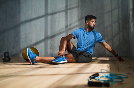 Young African American sportsman sitting on floor and doing stretching exercise indoors, workout training concept.