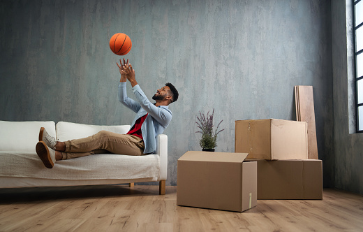 A happy young man with basketball celebrating moving home surrounded by packing boxes, new living concept.