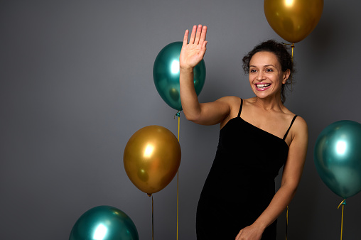Cheerful pretty woman dressed in elegant black evening dress stands against a gray wall background with golden and green metallic inflated balloons, waves hello with her hand, smiles with toothy smile