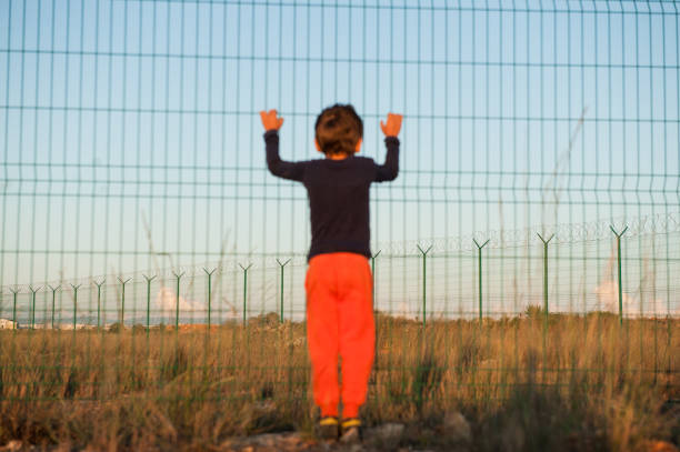 poor suffering little boy refugee in orange pants holding high fence on state border