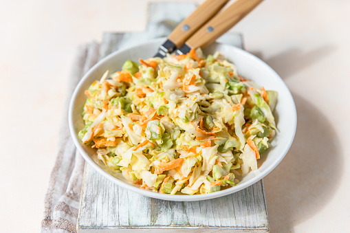 Coleslaw. Salad made of shredded white cabbage, grated carrot and rhubarb with orange mayonnaise dressing in white bowl, light concrete background.