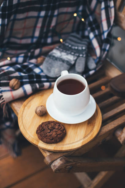 Still life with hot tea and chocolate cookies on wooden tray with warm blanket and festive lights on background. Cozy wooden terrace of a countryhouse. Slow life concept.