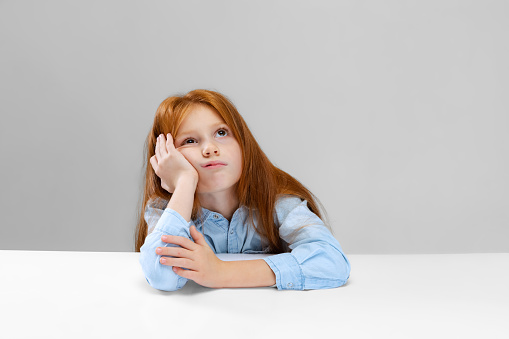 Portrait of little girl, pupil feeling bored, propping up cheeks isolated oer gray background Concept of childhood, family, happiness, education, emotions, facial expression. Copy space for ad