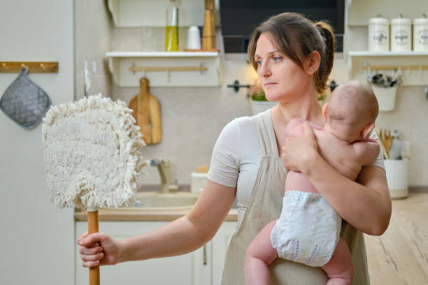 istock A woman holds an infant baby in her arms while cleaning in a home kitchen, problems with household chores after the birth of a child 1344908016