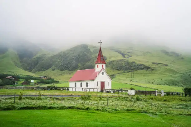 Photo of Typical Rural Icelandic Church with red roof in Vik region of Iceland. Green hills in mist in background