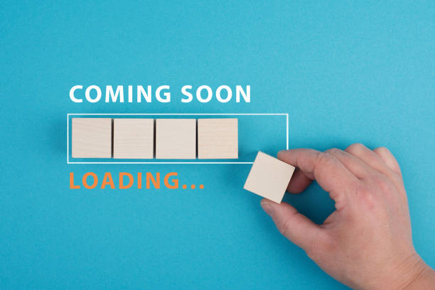The words coming soon are standing over a loading bar, hand puts the last wooden cube to the line, blue colored background stock photo