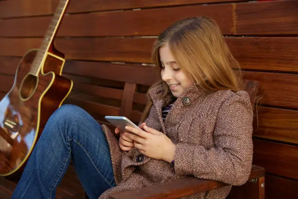 blond kid girl playing smartphone and guitar on wooden background